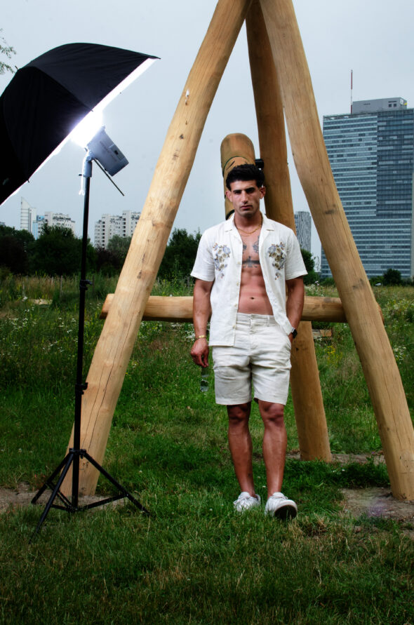 Male model wearing an open summer shirt and shorts in a nature environment, showing tattoos and a fit upper body. Flash lamp on the left side